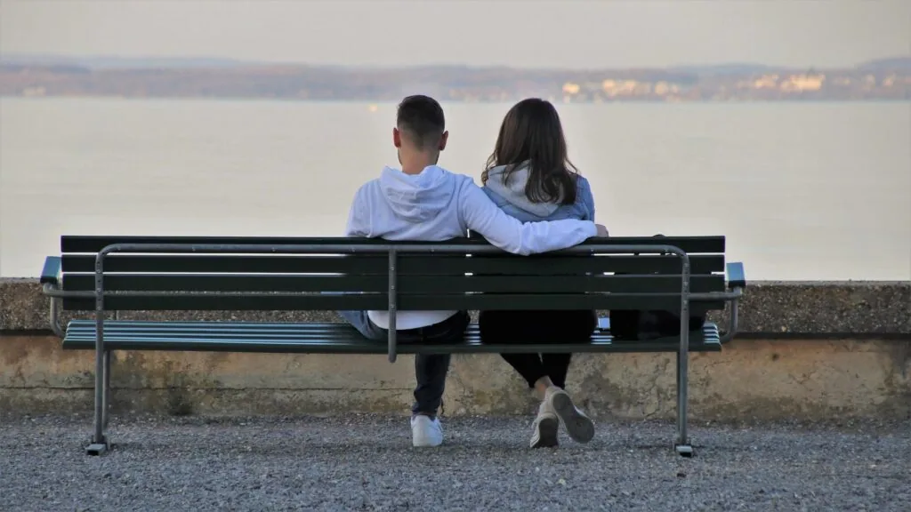 Two people sitting on a green bench facing a calm body of water with distant shoreline