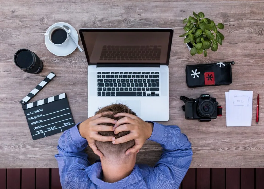 Person in blue shirt sitting at desk with laptop, coffee, film clapper, camera, potted plant, and office supplies viewed from above