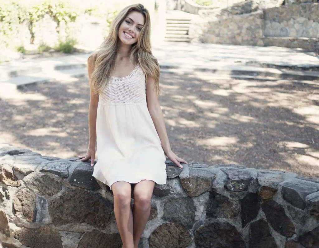 Woman in white sleeveless dress sitting on stone wall in sunlit courtyard