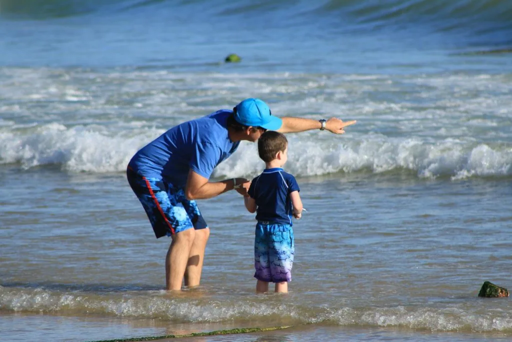 Adult wearing blue cap and child in dark shirt playing together in shallow ocean waves at the beach
