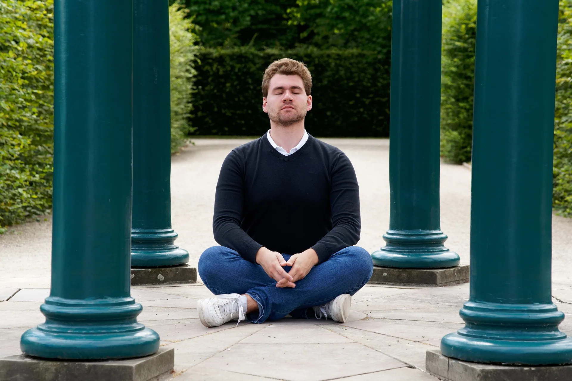 Man sitting cross-legged between two teal columns in a garden setting